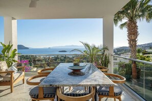Ein Balkon mit einem Marmortisch, Holzstühlen und Blick auf das Meer und ferne Berge.
