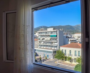 A window view of a multi-story building with balconies, a clear sky, and distant mountains. - Cozy flat on the main square, 10min to the sea (Kalamata)