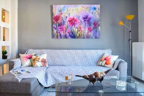 A modern living room with a grey sofa, floral cushions, a glass coffee table, and a colorful abstract painting on the wall.