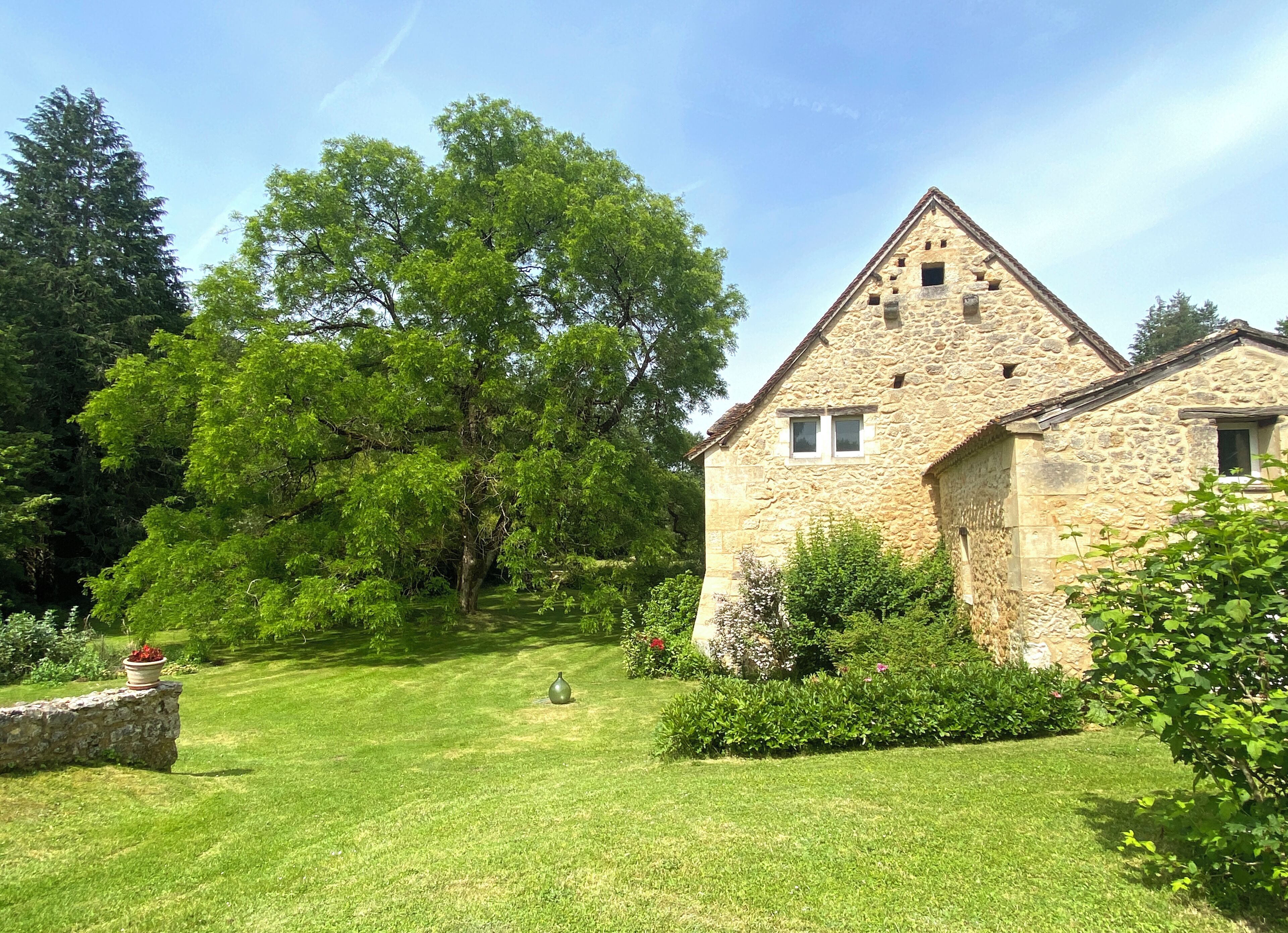 Un bâtiment en pierre doté d’un toit triangulaire, entouré d’une pelouse bien entretenue et de divers arbustes.