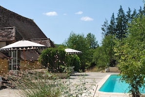 A swimming pool with a tiled deck, surrounded by greenery and a building with a sloped roof. - Gîte du verger avec piscine dans un domaine périgourdin (Saint-Georges-de-Montclard)