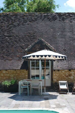 Un bâtiment en pierre avec un toit en tuiles, un petit patio avec une table et des chaises, et une piscine.