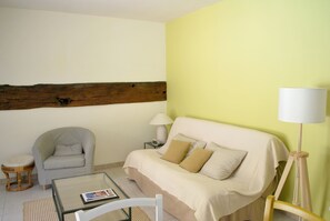 A room with a sofa, a chair, a small table with a magazine, a lamp, and a wooden beam on the wall. - Gîte du verger avec piscine dans un domaine périgourdin (Saint-Georges-de-Montclard)