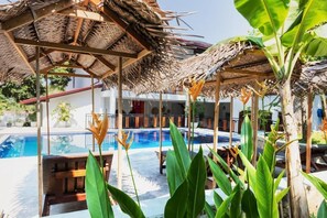 Un área de piscina con una estructura de techo de paja, rodeada de abundante vegetación y una piscina azul cristalina.