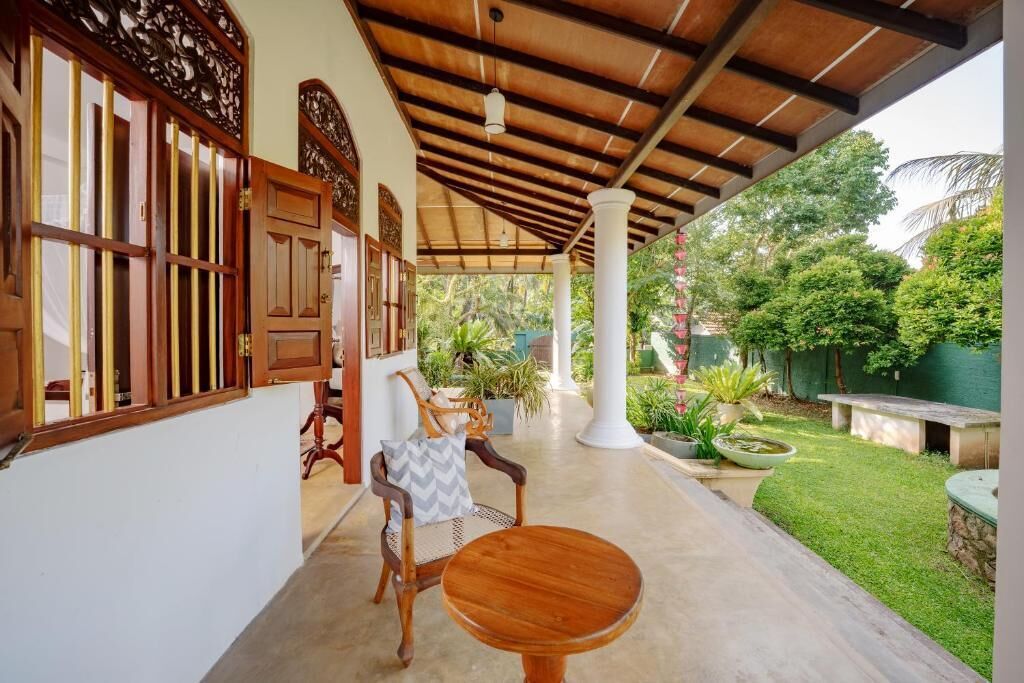Un espacio al aire libre cubierto con una mesa y sillas de madera, un pilar blanco, y un jardín con plantas y un pequeño estanque.