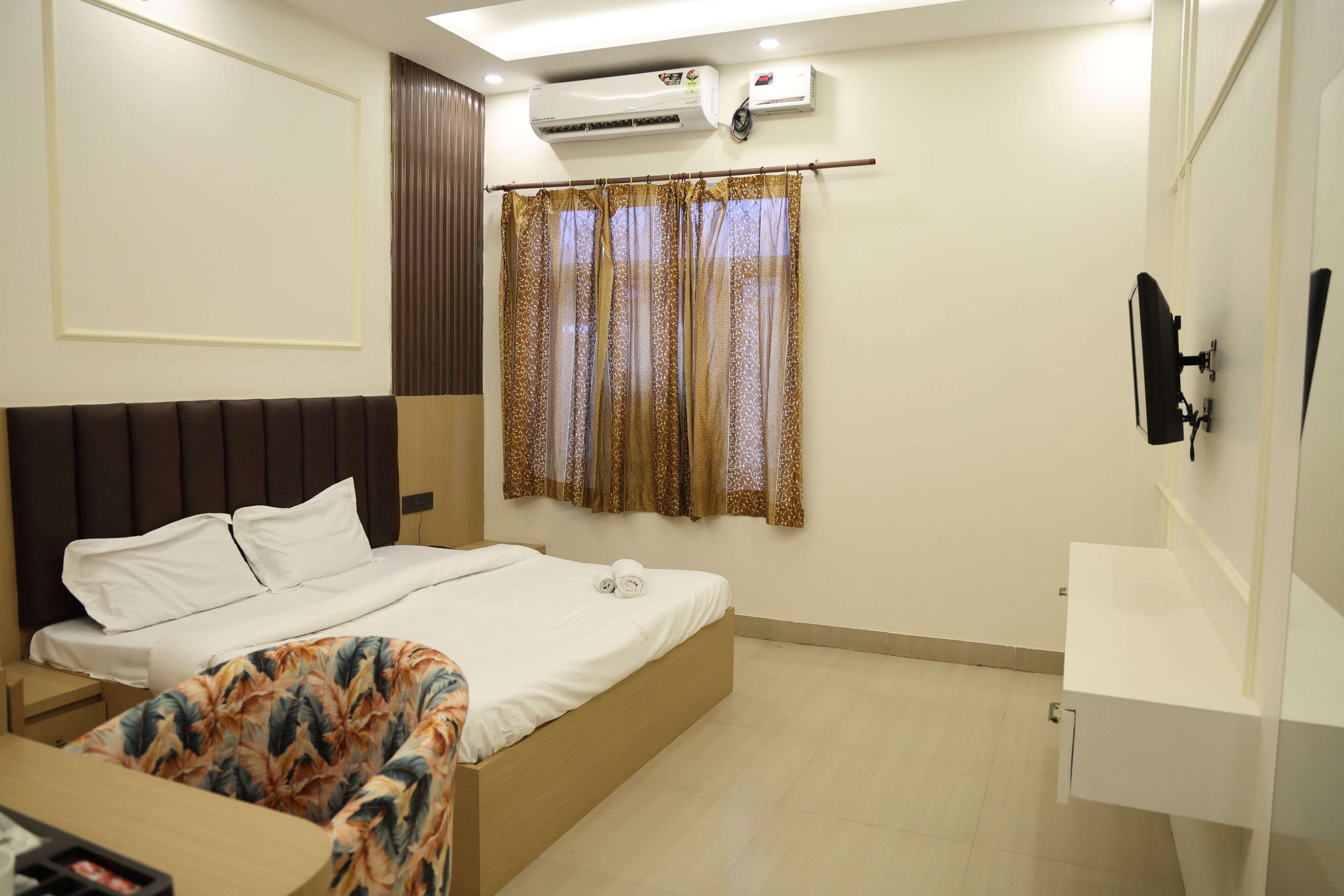 A hotel room with a bed, a desk, a TV mounted on the wall, and an air conditioning unit.