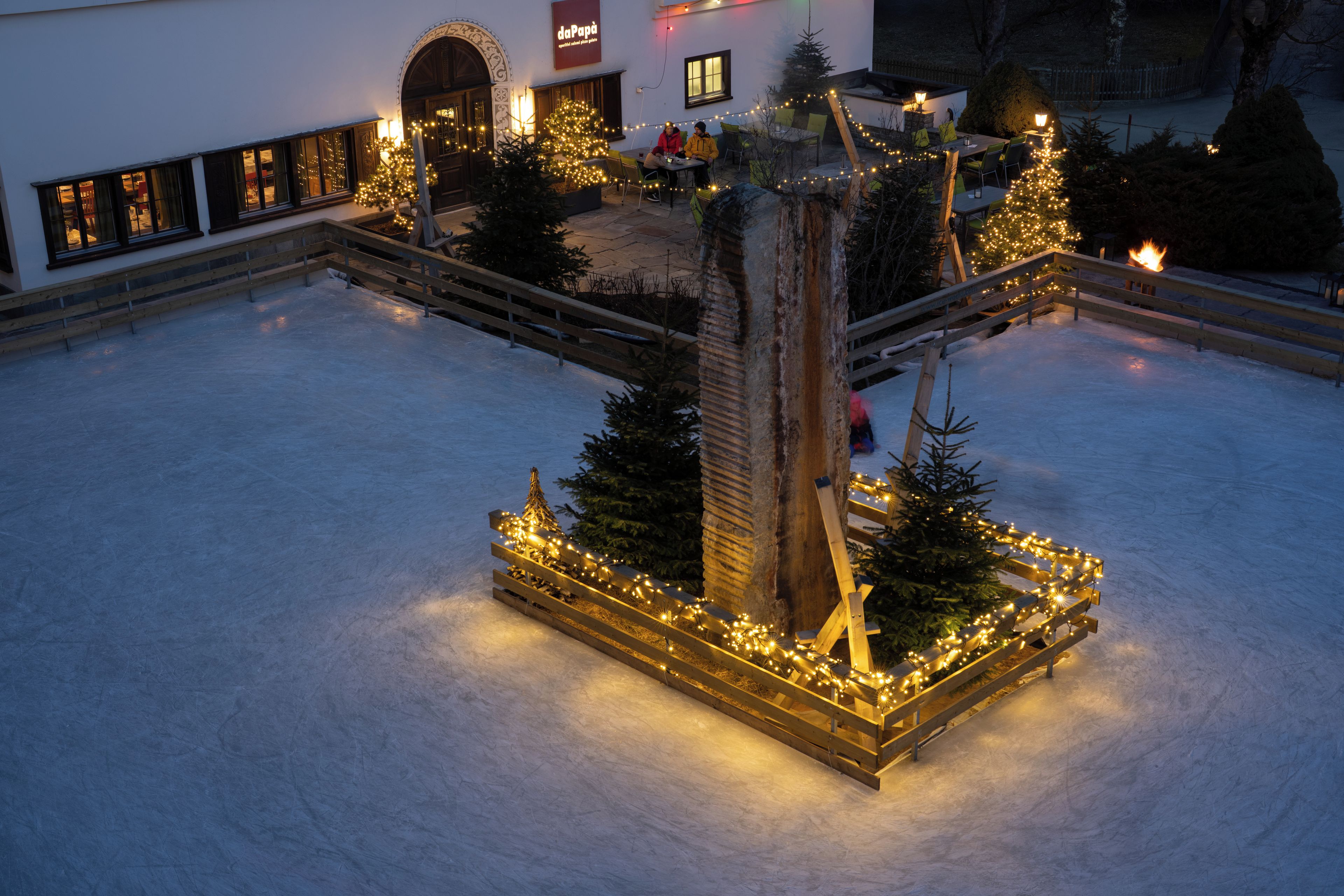 Ein verschneiter Innenhof mit einer zentralen Holzkonstruktion, verziert mit Lichtern, umgeben von Weihnachtsbäumen und einer Sitzgelegenheit.