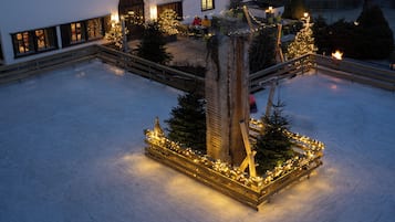 Ein verschneiter Innenhof mit einer zentralen Holzkonstruktion, verziert mit Lichtern, umgeben von Weihnachtsbäumen und einer Sitzgelegenheit.