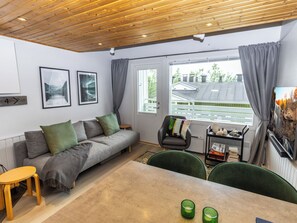Ett modernt vardagsrum med ett träpanelat tak, en grå soffa med gröna kuddar, ett matbord med gröna stolar och ett fönster med utsikt över ett hus och träd.