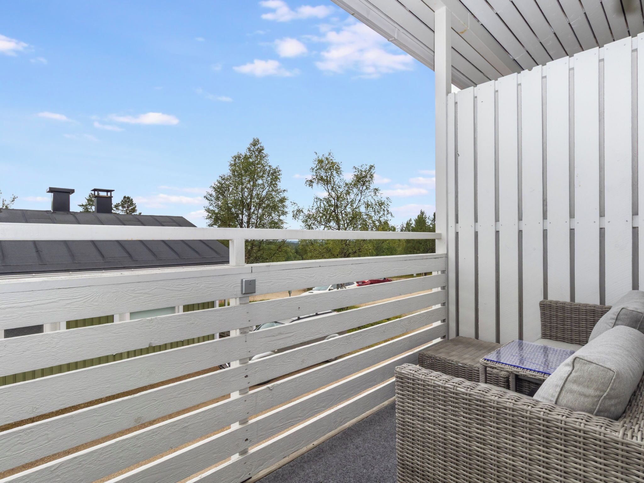 A covered outdoor seating area with a white railing, wicker furniture, and a view of the sky and trees.