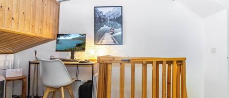 A room with a wooden desk, a computer, a chair, a bed with a wooden frame, a window, and a wall-mounted picture.