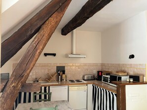 A compact kitchen with a dishwasher, stove, and microwave. A wooden beam structure is visible, and a small dining table with chairs is placed under the beam. - Gîte du potager avec piscine dans un domaine périgourdin (Saint-Georges-de-Montclard)