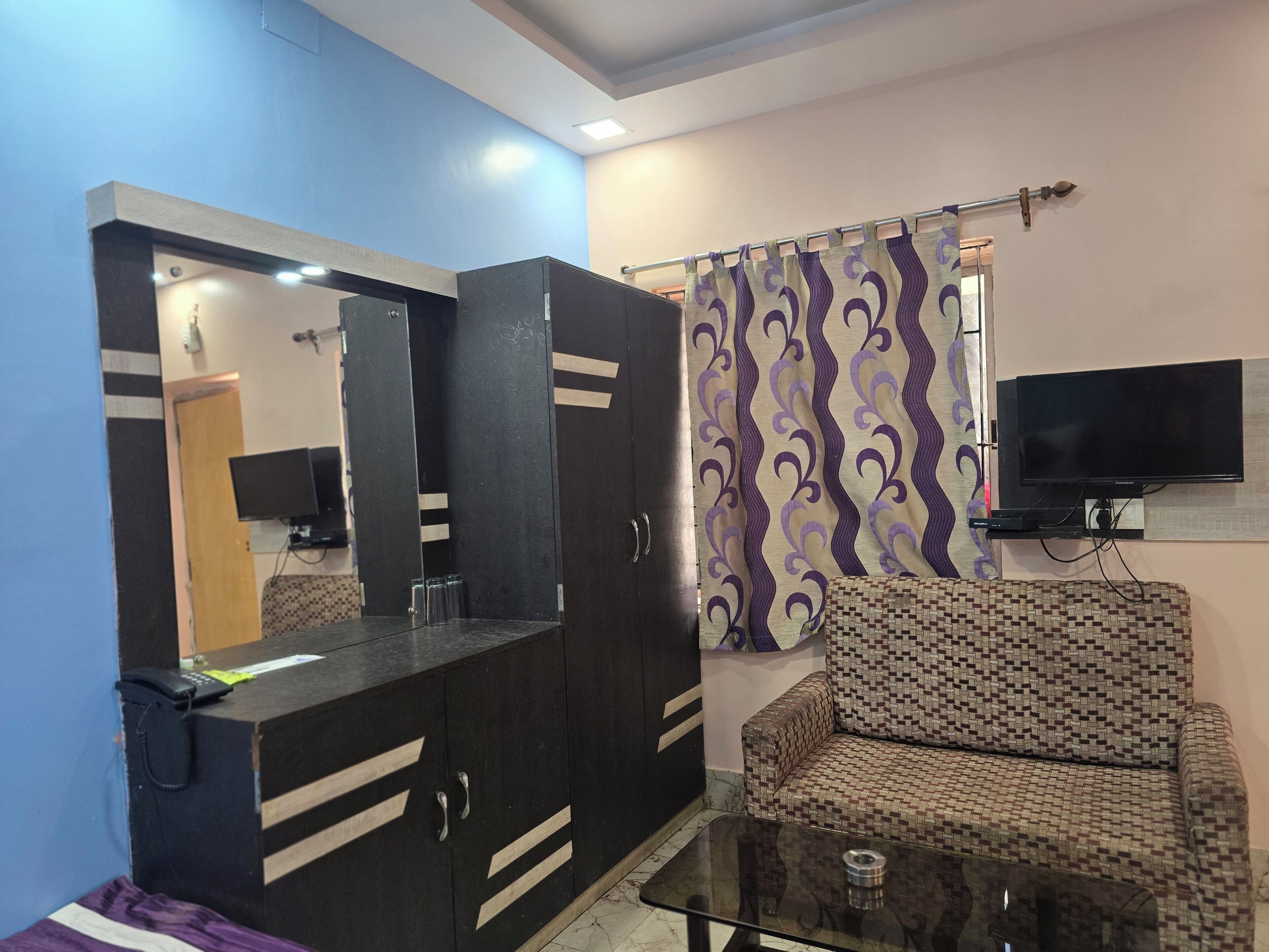 A room with a sofa, a television mounted on the wall, a cabinet with a mirror, and a patterned curtain.