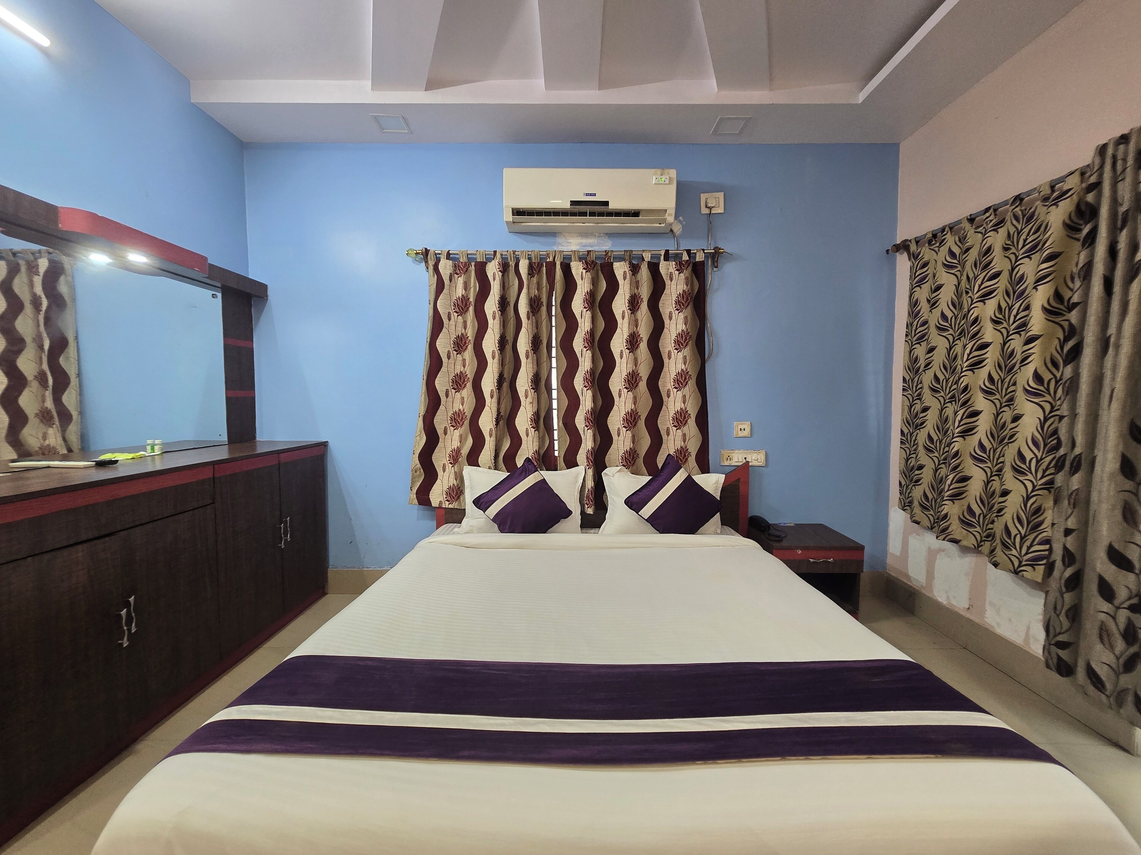 A hotel room with a bed, a wardrobe, an air conditioning unit, and patterned curtains.