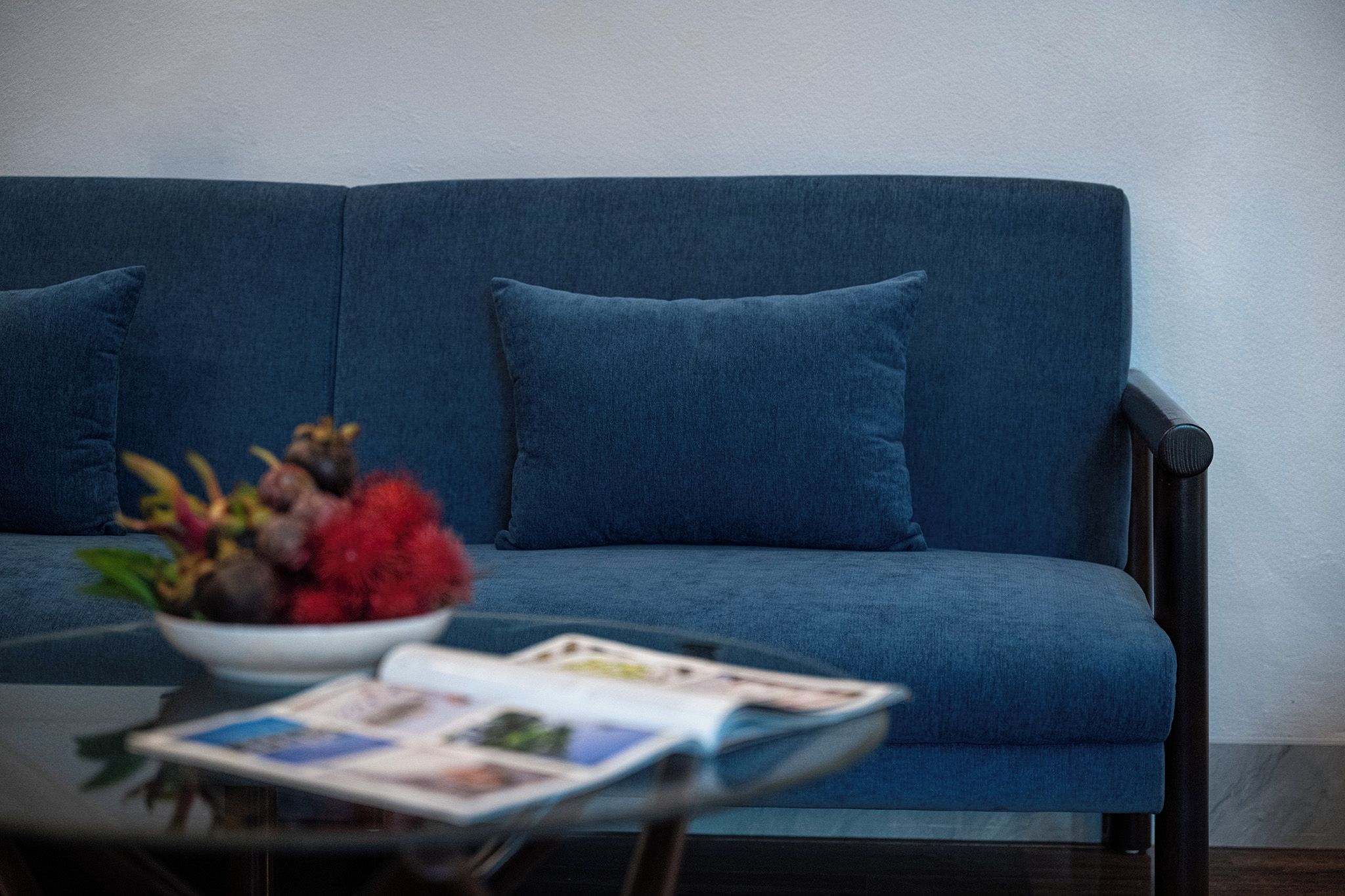 En blå soffa med en skål med frukt och en tidning på ett bord med glasskiva.