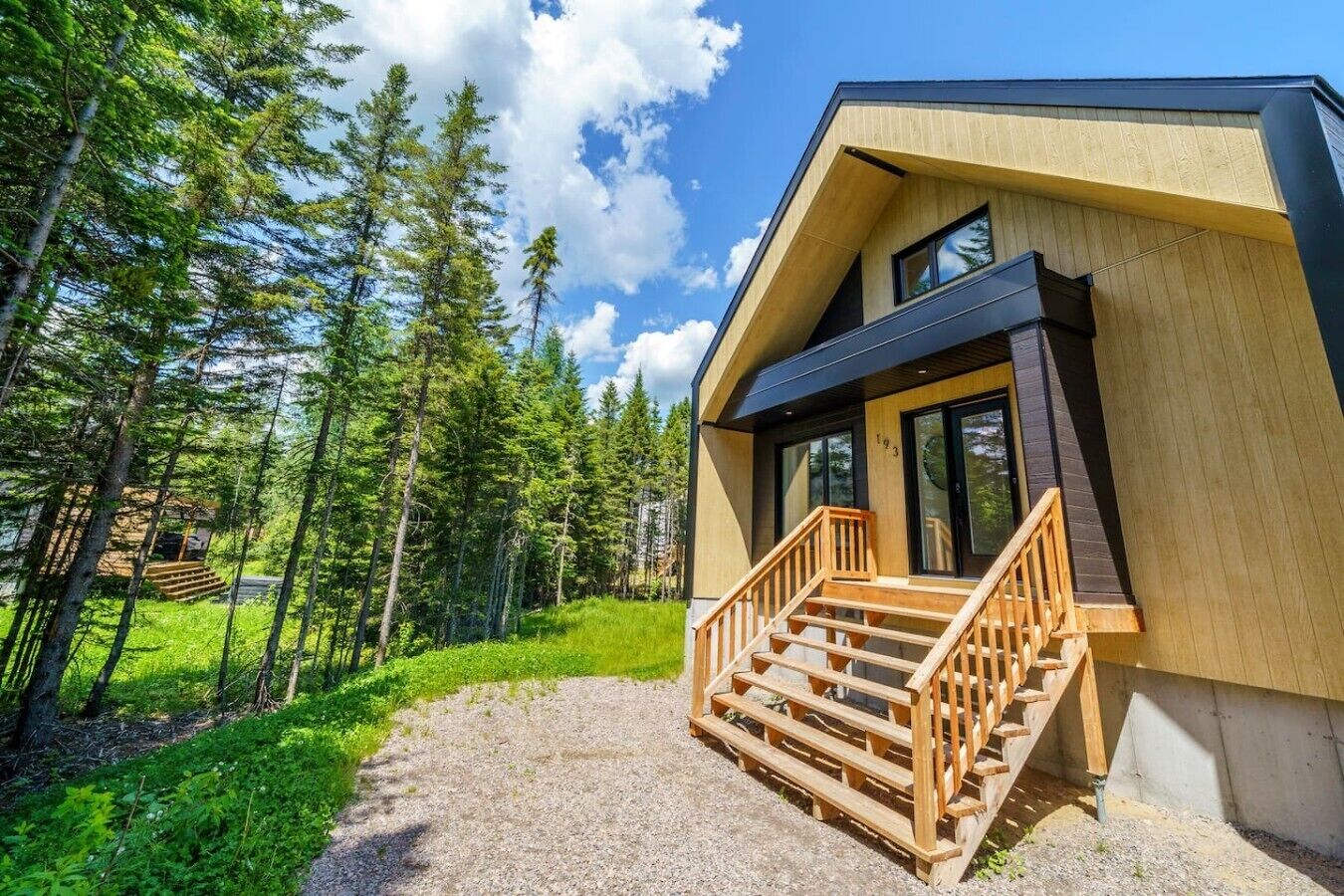 A modern wooden cabin with a wooden staircase leading to a glass door, surrounded by a forest.