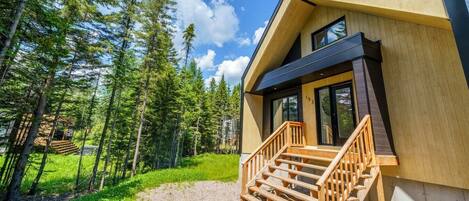 A modern wooden cabin with a wooden staircase leading to a glass door, surrounded by a forest.