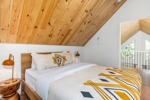 Ein Schlafzimmer mit schrägen Wänden, einem Bett mit gemustertem Überwurf, Nachttischlampen und Blick ins Freie.