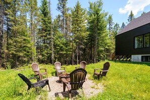 Ein modernes Haus mit einer Terrasse, umgeben von einem Wald. Draußen stehen Stühle und ein Tisch.