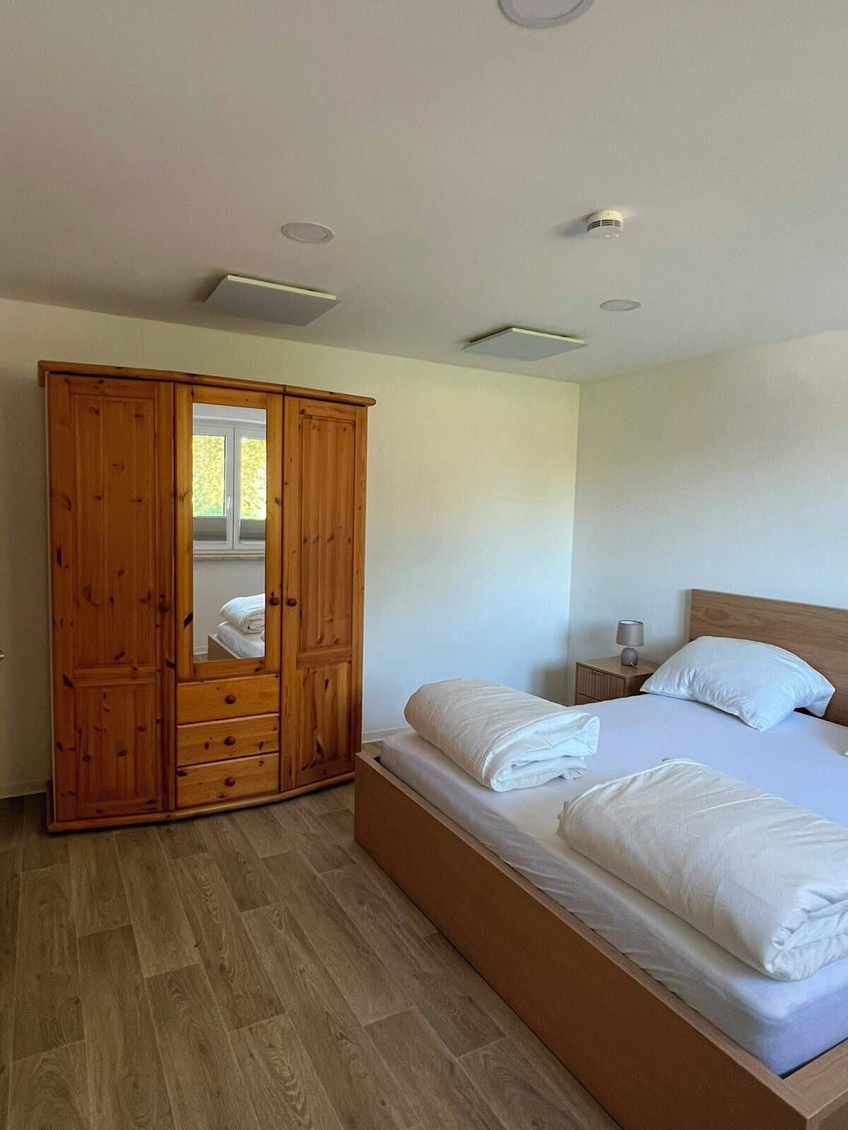 Une chambre avec une armoire en bois, un lit avec du linge de lit blanc et une table de chevet avec une lampe.