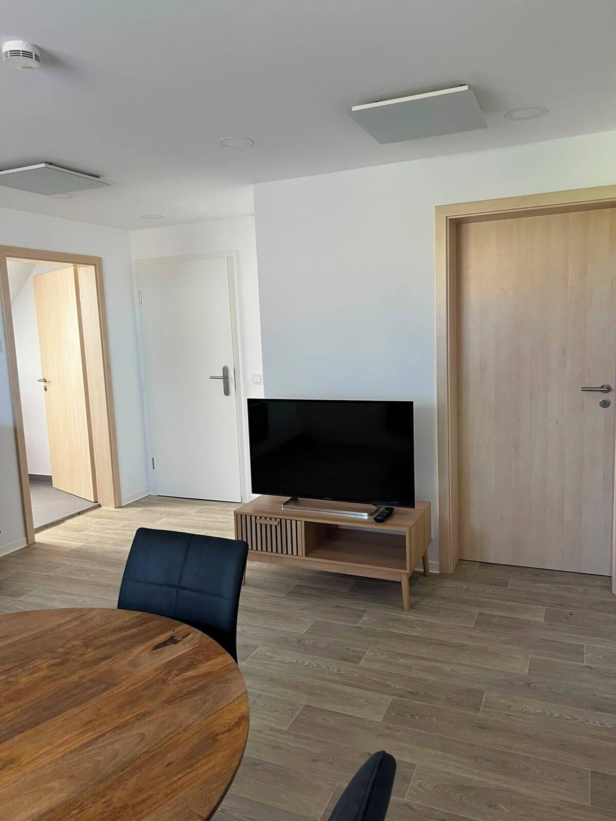 Ein modernes Wohnzimmer mit einem Flachbildfernseher auf einem Holzboden, einem runden Holztisch und einem schwarzen Sessel. Es gibt zwei Türen und eine Deckenleuchte.
