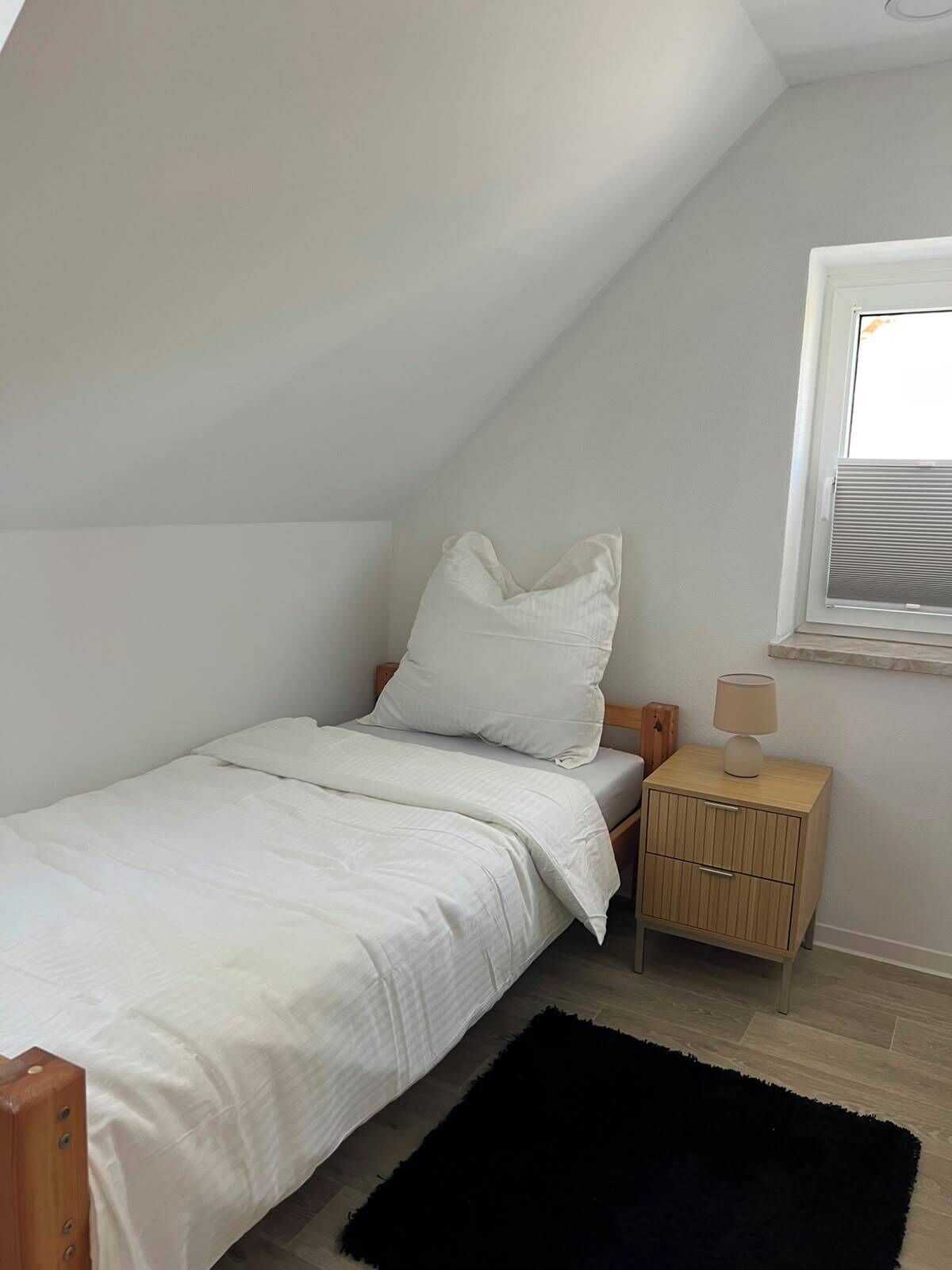 A small, single-bed room with a sloped ceiling, a bedside table with a lamp, and a window with blinds.