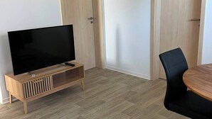 A room with a flat-screen TV on a wooden stand, a dining table with a black chair, and two wooden doors.