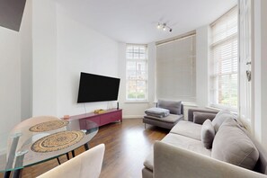 A modern living room with a flat-screen TV, a glass coffee table, a sofa, and a dining table with a patterned placemat. - Amazing 1BR Flat w/ Communal Garden and Parking (London)