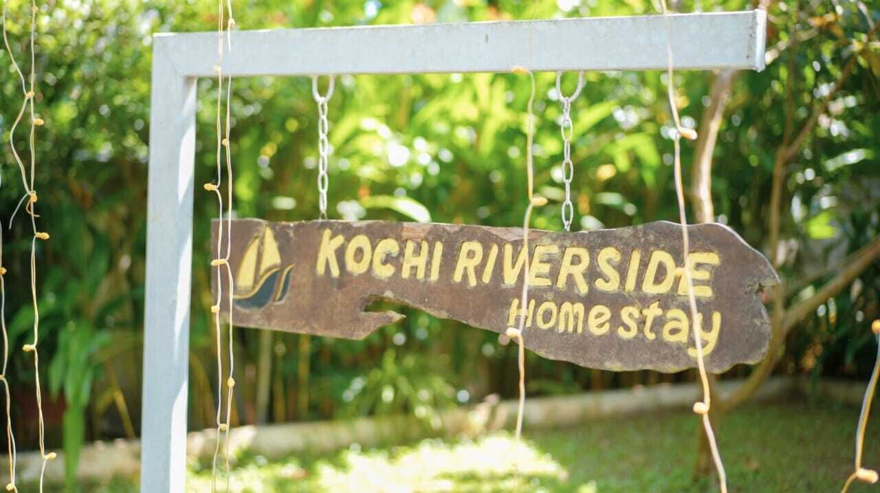 A wooden sign with the text 'KOCHI RIVERSIDE Home stay' hanging from a white structure.