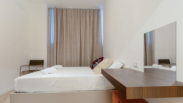 Una habitación de hotel moderna con una cama, una mesita de noche de madera, un espejo grande y una ventana con cortinas.