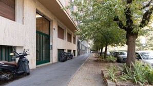Una estrecha calle urbana con scooters y automóviles estacionados, una acera arbolada y un edificio con un portón verde.