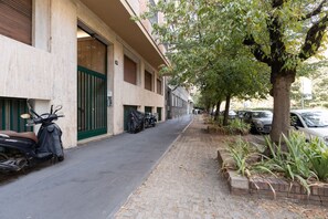 Eine schmale Stadtstraße mit geparkten Rollern und Autos, einem von Bäumen gesäumten Bürgersteig und einem Gebäude mit einem grünen Tor.