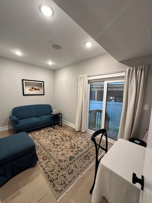 Ein Wohnzimmer mit blauem Sofa, einem kleinen Tisch, einem gemusterten Teppich und einem Balkon mit Vorhängen.