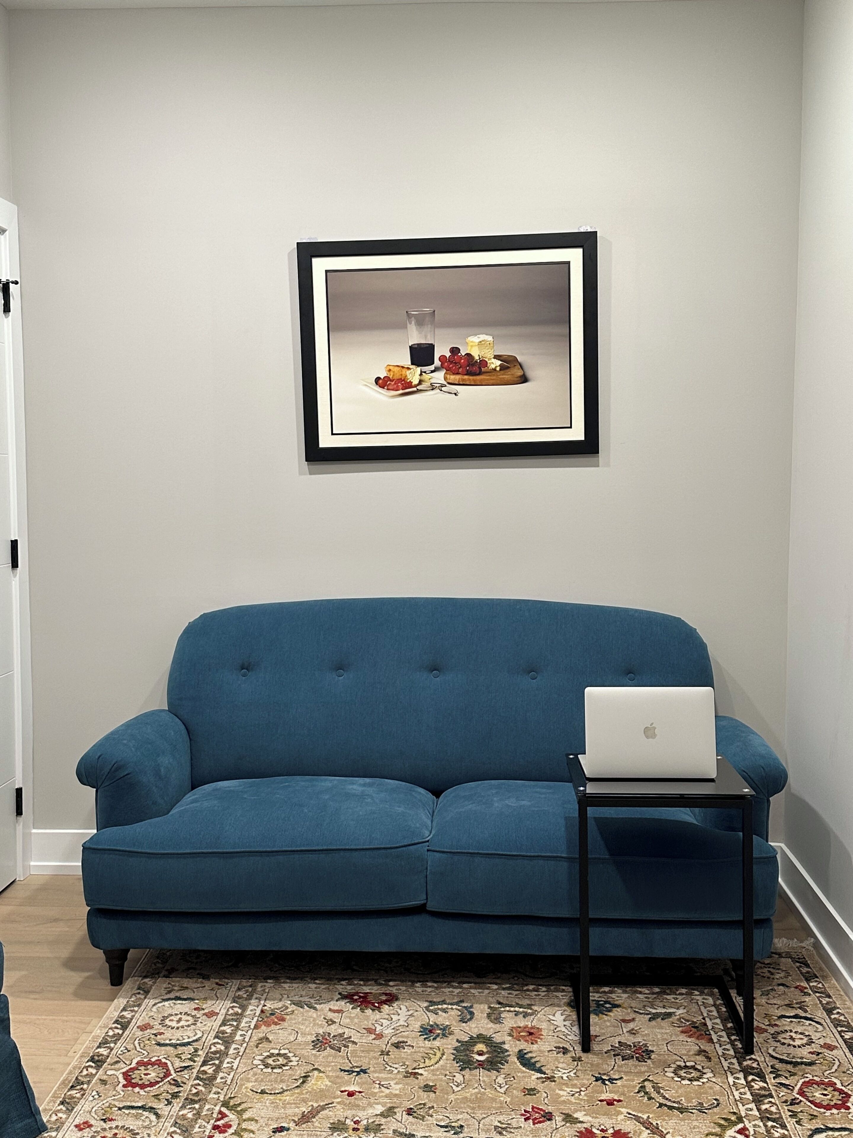 Une pièce avec un canapé bleu, une petite table d’appoint noire sur laquelle se trouve un ordinateur portable, un tableau encadré au mur et un tapis à motifs.