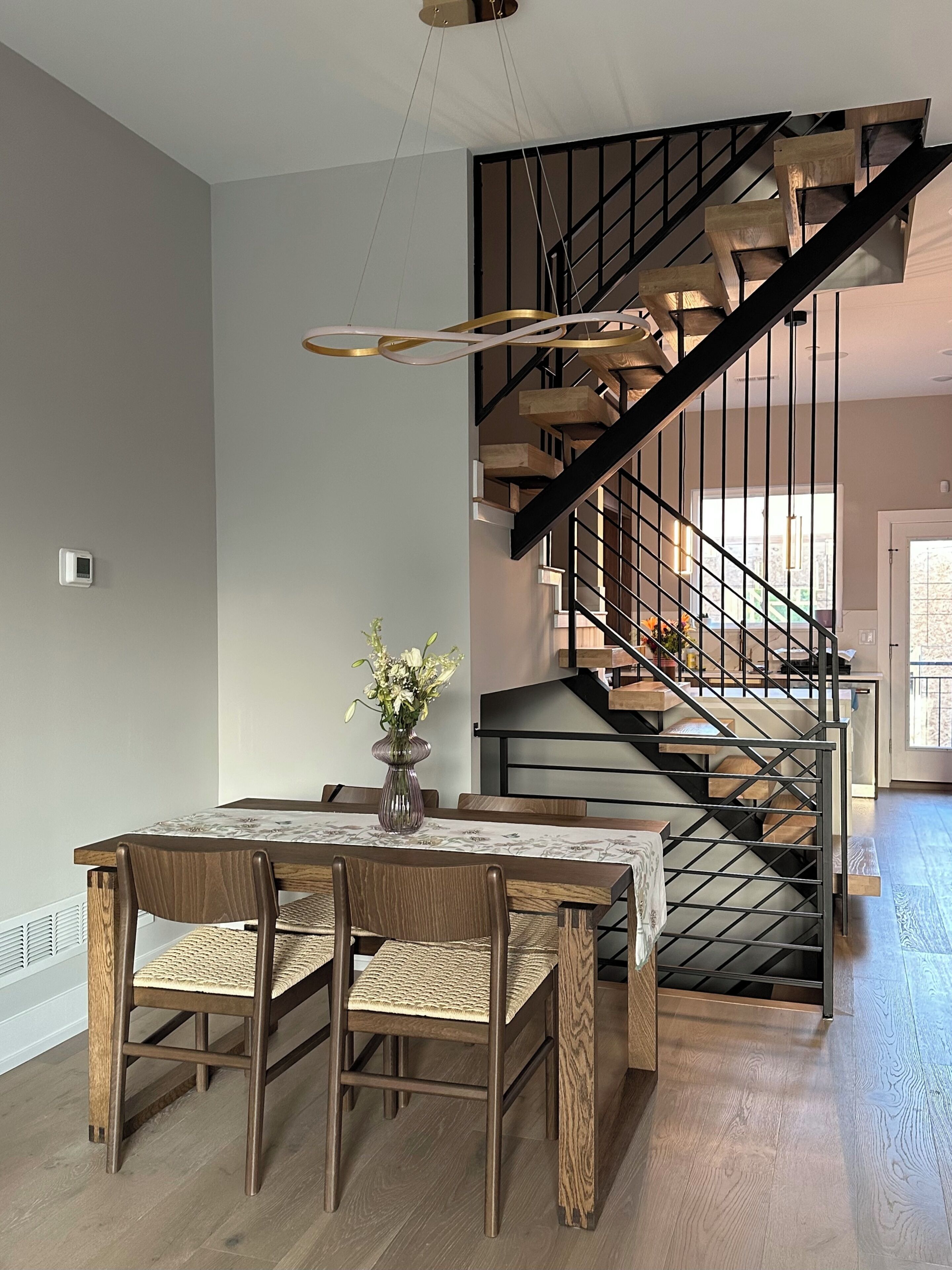 A dining area with a wooden table and chairs, a vase with flowers, and a staircase with wooden steps and metal railings.
