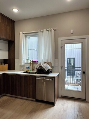 A modern kitchen with a dishwasher, wooden cabinets, a window with curtains, and a door leading to a balcony. - Modern South Philly Townhome. Rooftop. Skyline Views. Home Office.  Gym (Philadelphia)