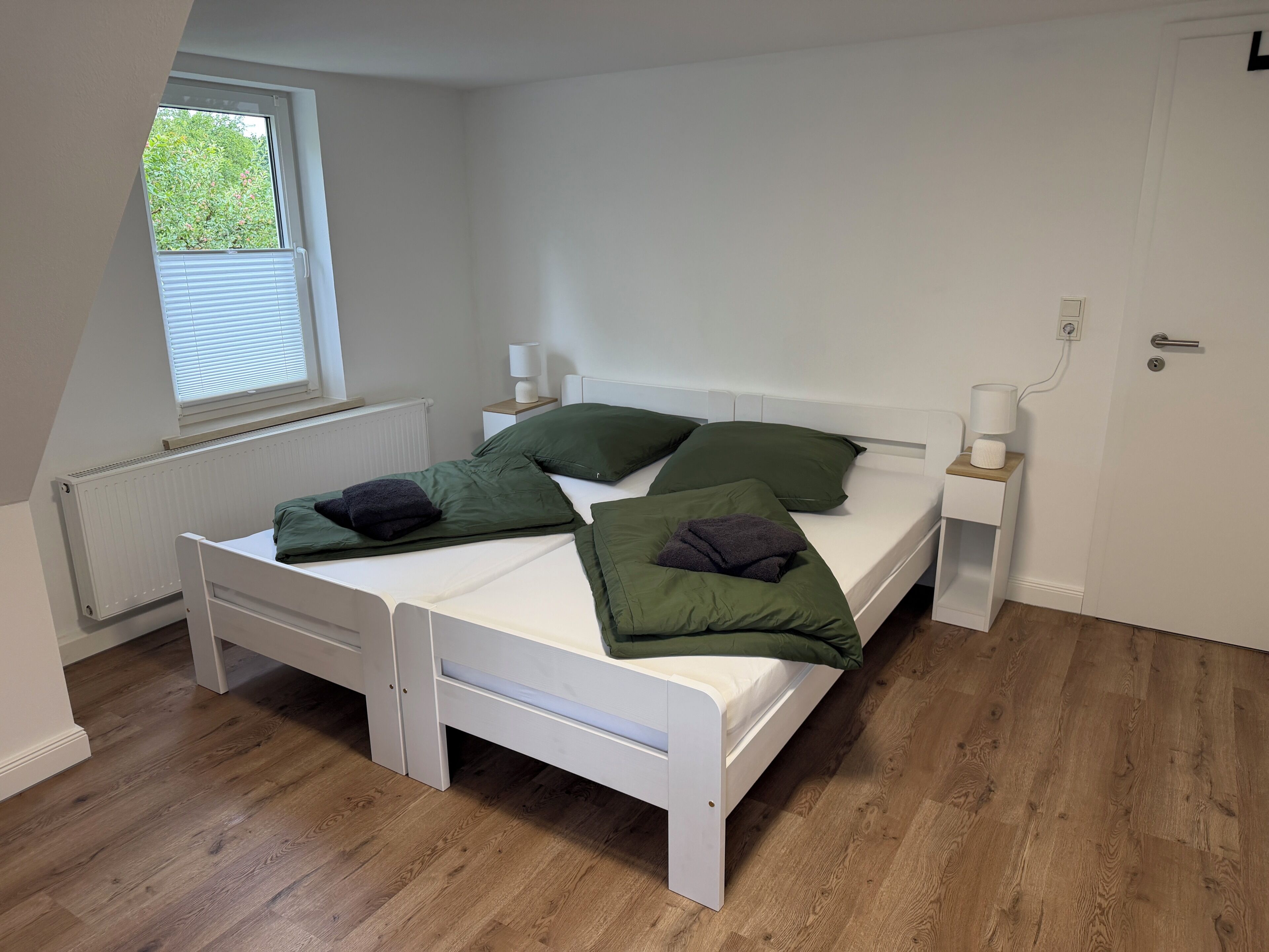 A bedroom with a white bed, green pillows, a bedside table with a lamp, and a window with blinds.