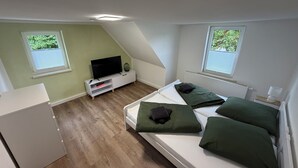 Ein kleines, modernes Schlafzimmer mit Mansardendecke, einem Flachbildfernseher auf einer weißen Konsole, einem Bett mit grünen Kissen und zwei Fenstern mit Jalousien.