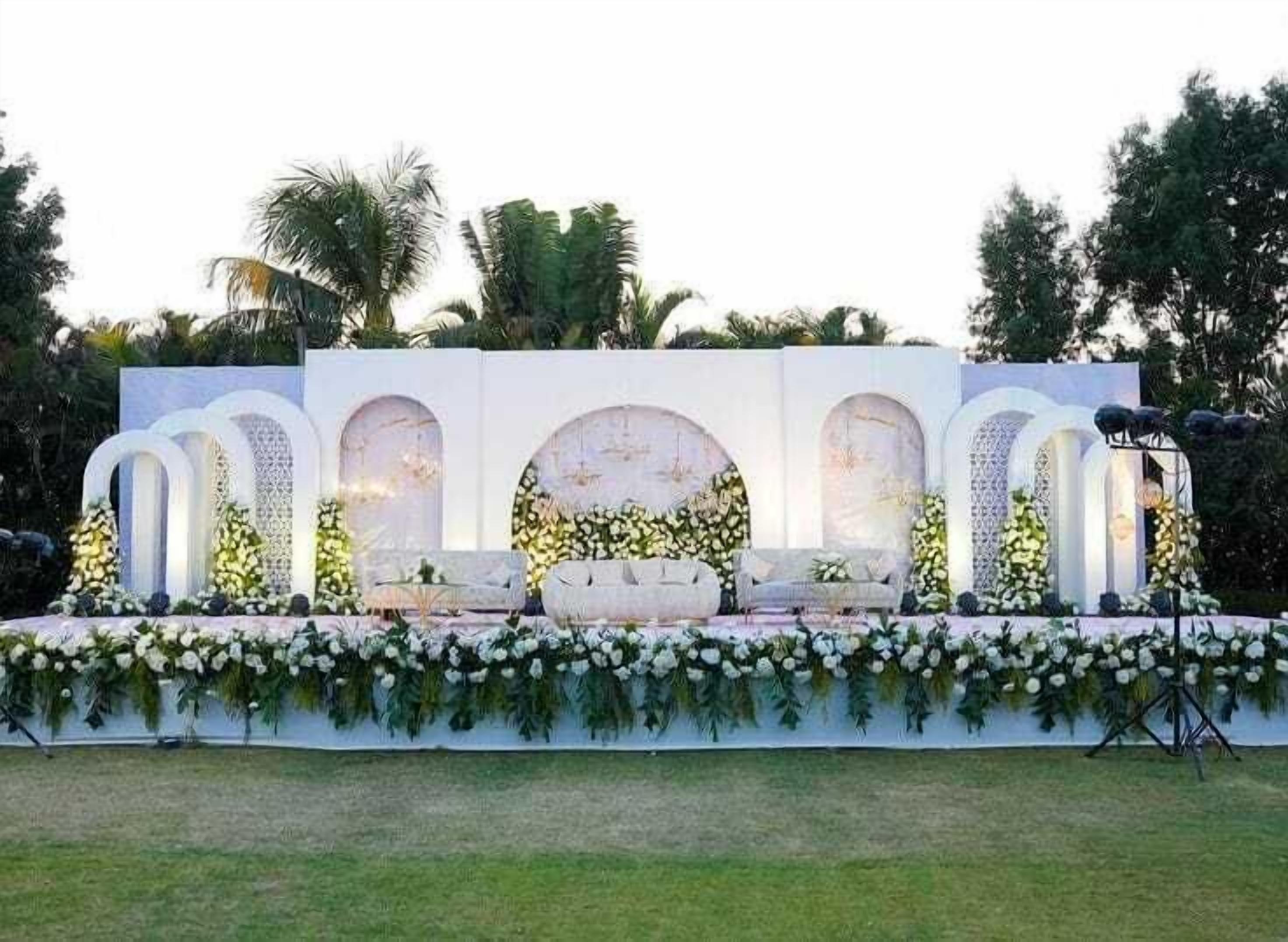 An outdoor stage with a white backdrop, floral arrangements, and seating.