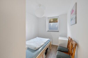 Una habitación pequeña con cama individual, ventana, mesita de noche y una silla.