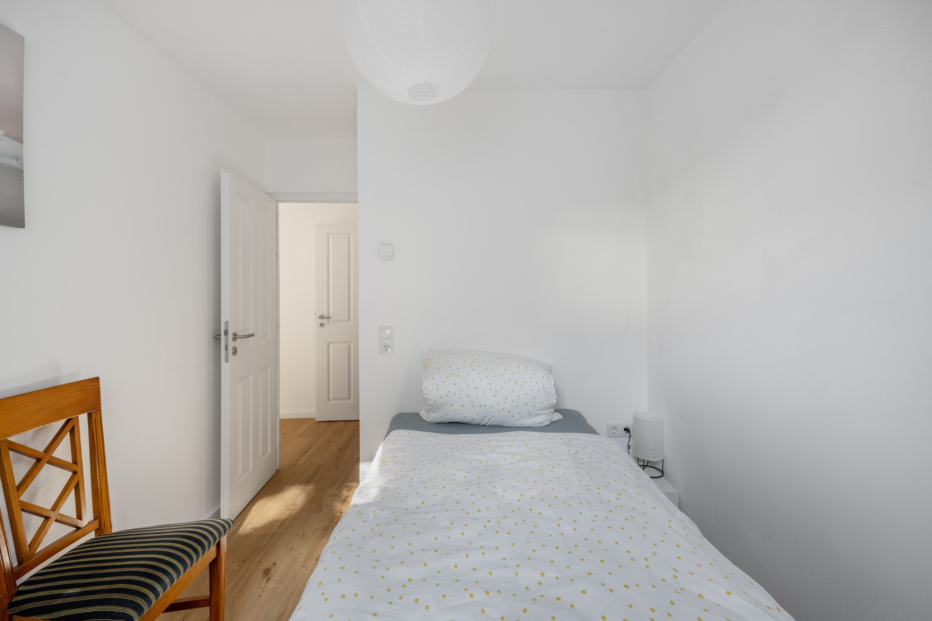 Une petite pièce étroite, avec un lit, une chaise et une porte donnant sur une autre pièce.