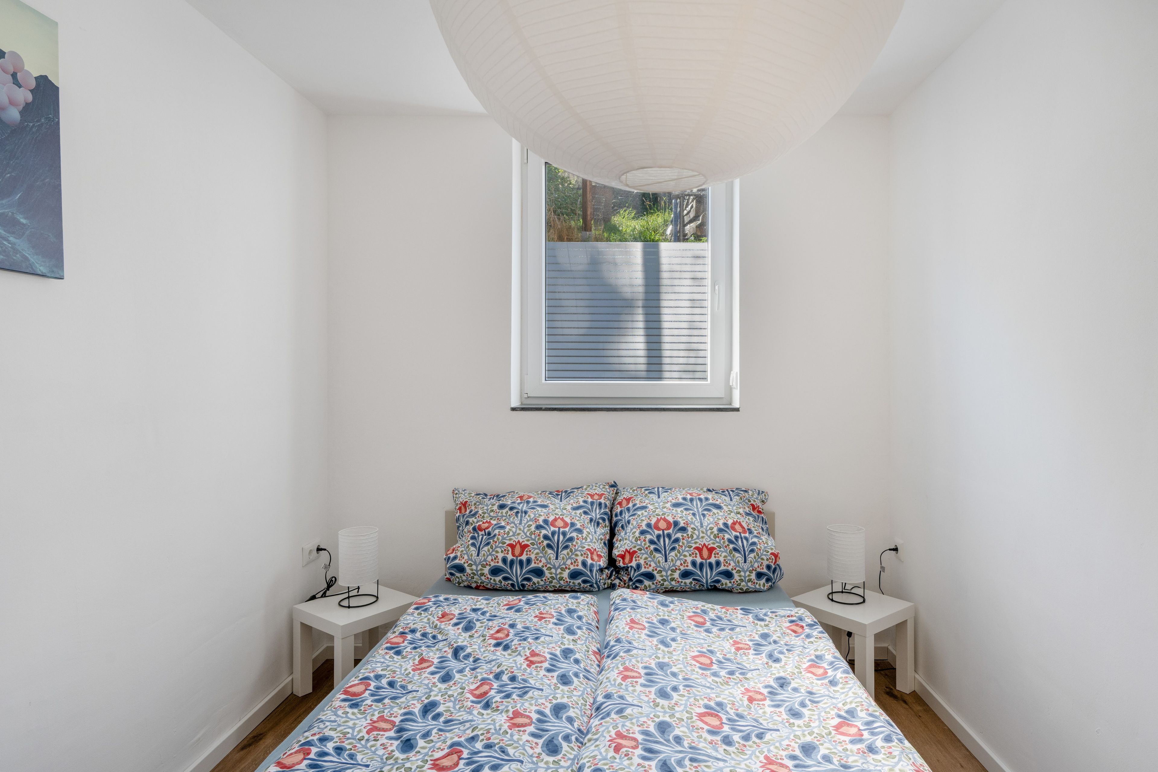 Ein Schlafzimmer mit einem Bett, zwei Nachttischen, einem Fenster mit Jalousien und einer Wandleuchte.