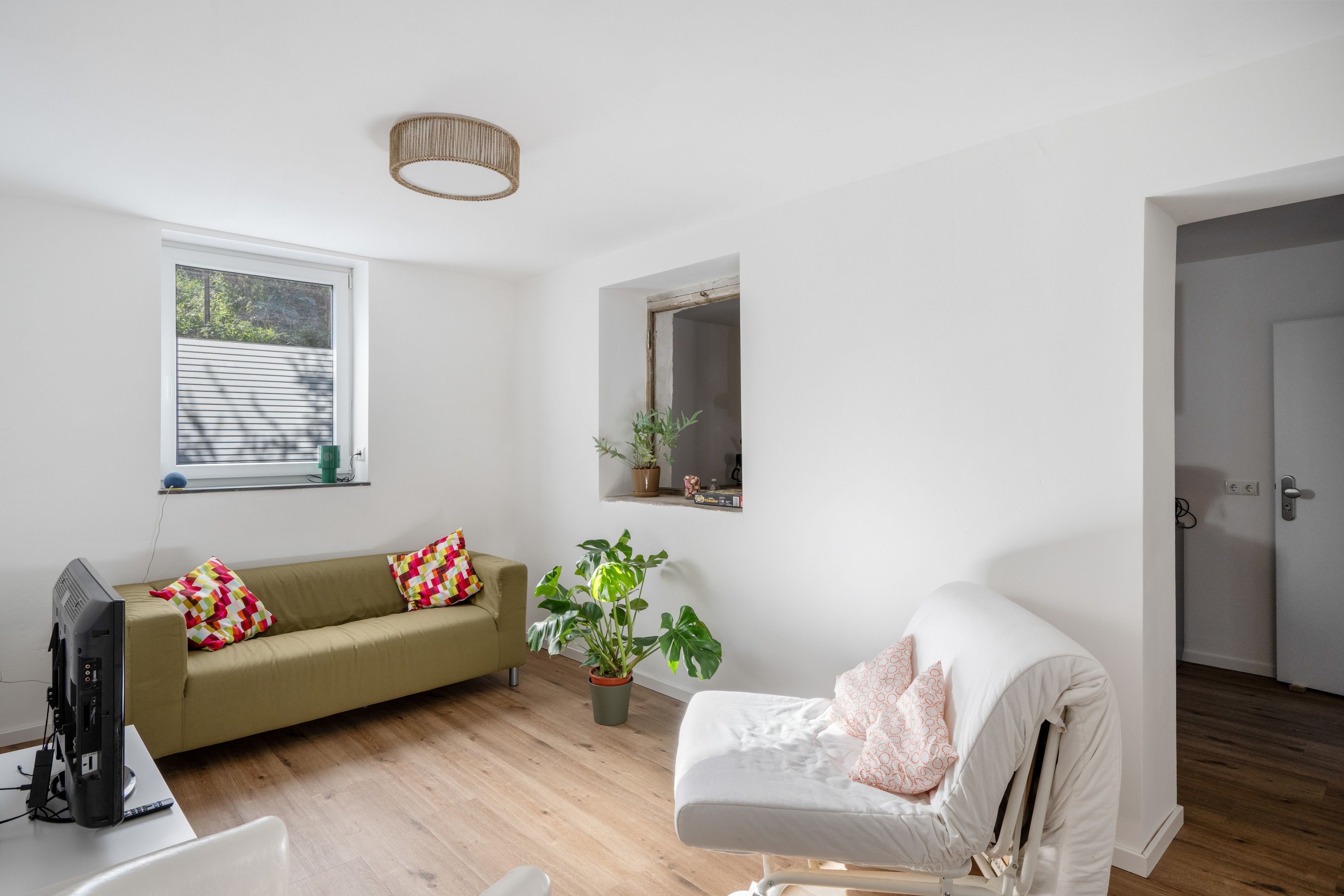 Un salon comprenant un canapé, un fauteuil, un téléviseur, une plante en pot et une fenêtre avec des stores.