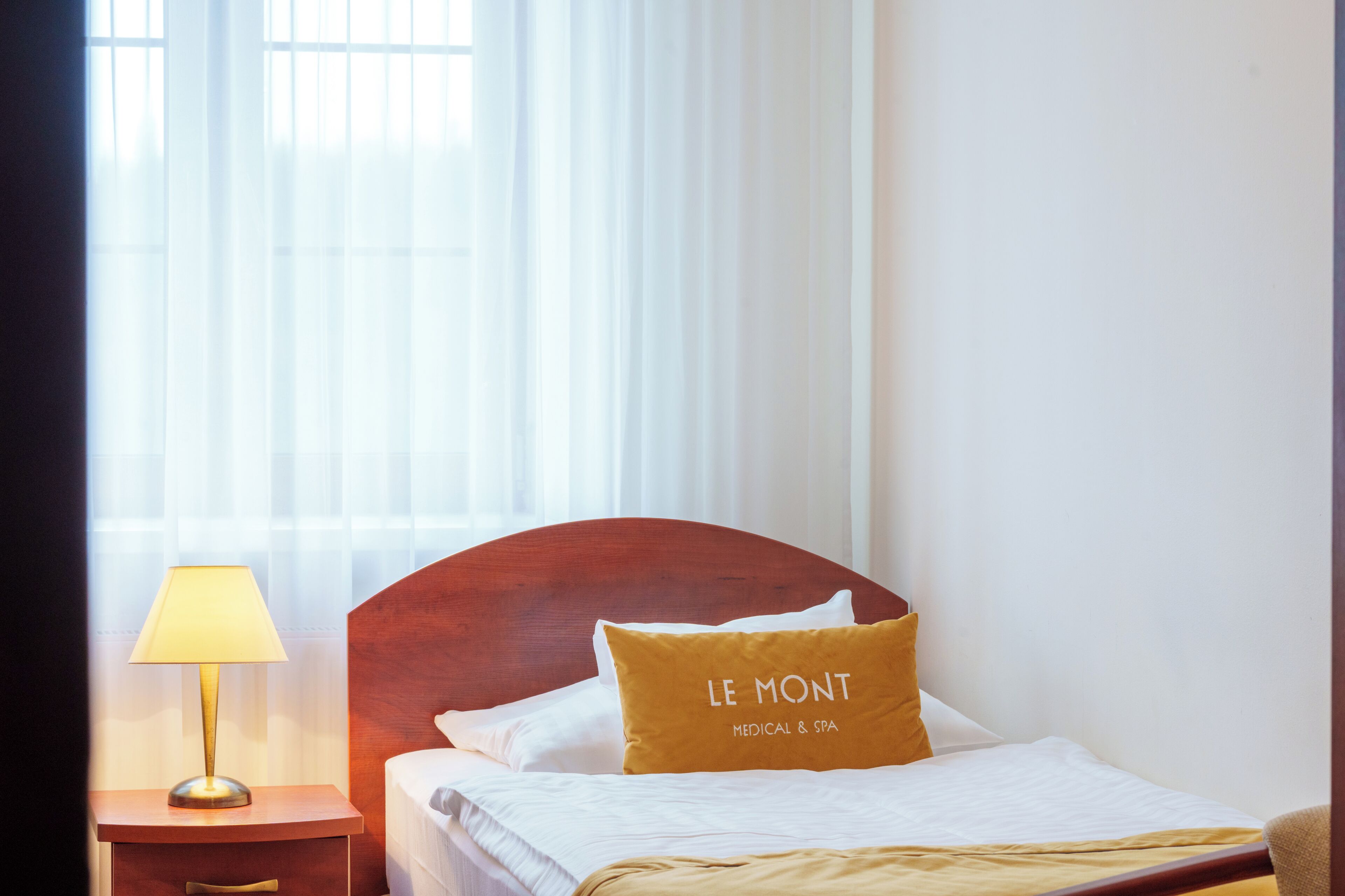 Quarto de hotel com uma cama, mesa de cabeceira com luminária e janela com cortinas leves.