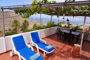 Una terraza con dos tumbonas, una mesa y sillas, con vistas a un paisaje costero con palmeras y vista al mar.