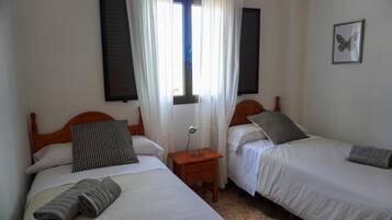Een kamer met twee bedden, elk met een houten hoofdeinde, witte beddengoed en een gestreept kussen. Daartussen bevindt zich een nachtkastje met een lamp, en er is een raam met gordijnen.