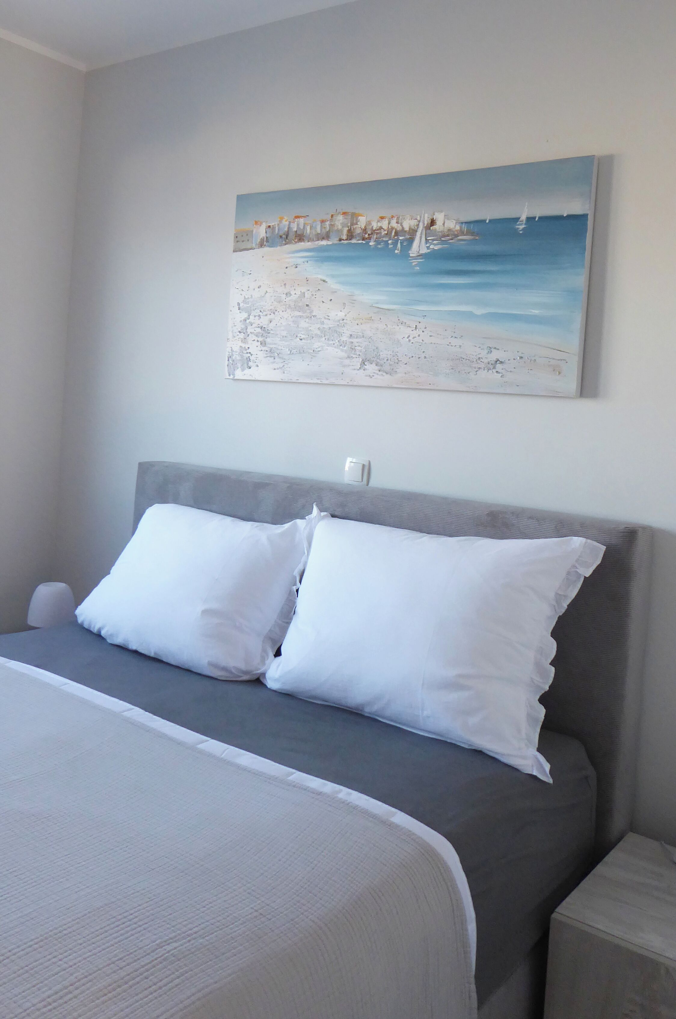 Une chambre à coucher avec un lit, deux oreillers, une table de chevet et un tableau représentant une scène de plage accroché au mur.