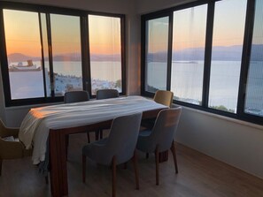 A room with a bed, two chairs, and large windows with a view of a body of water and distant hills.