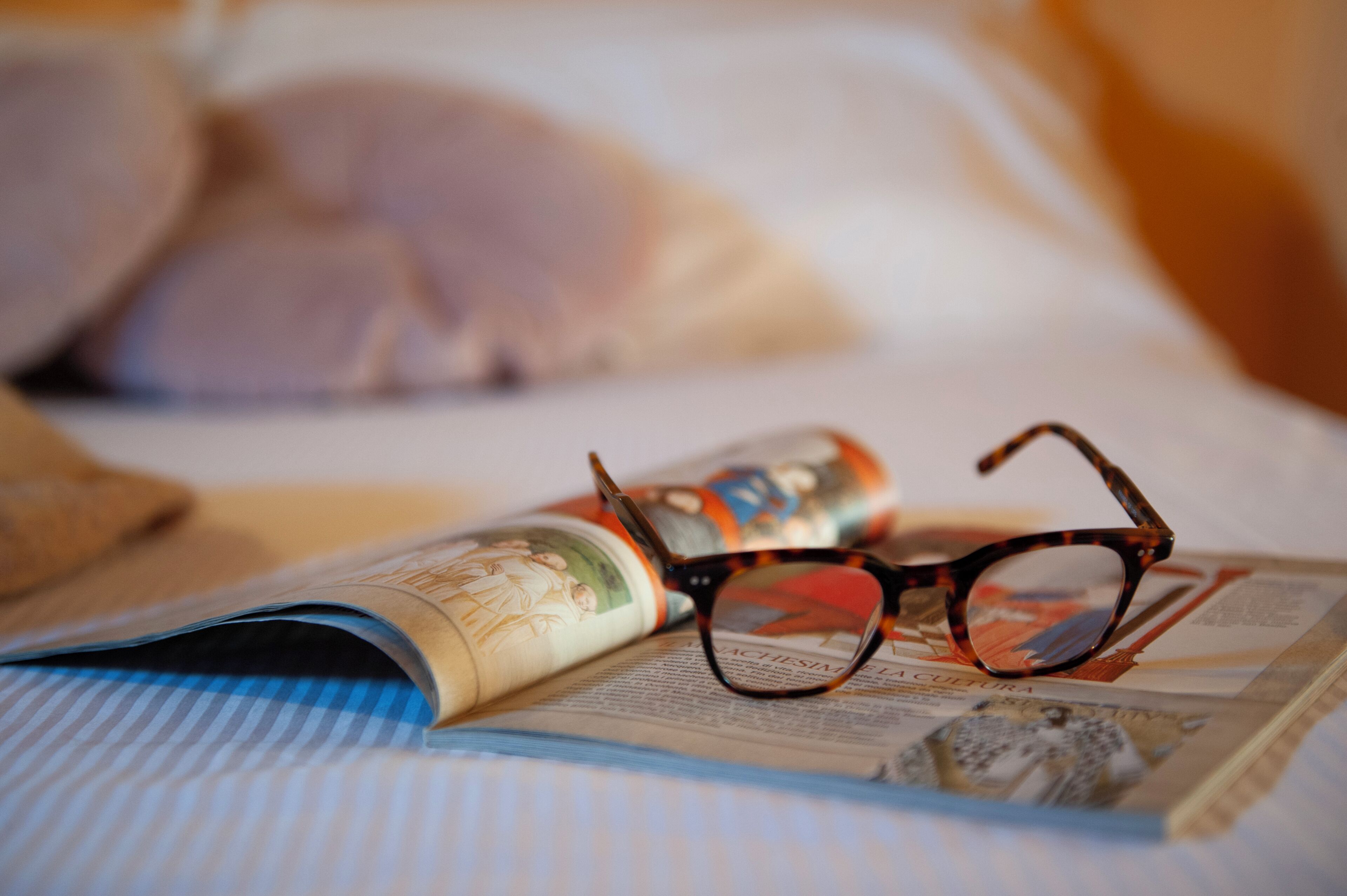 A pair of glasses on a magazine, a pillow, and a striped blanket on a bed.