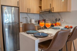 Une cuisine moderne avec un réfrigérateur en acier inoxydable, un plan de travail en marbre et des armoires en bois. On peut voir deux assiettes bleues, deux verres de jus d’orange et un saladier de fruits sur le plan de travail.